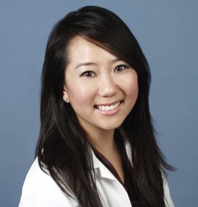 Professional headshot of smiling Asian woman in white blazer against blue background.