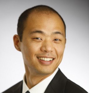 Professional headshot of an Asian man in business attire smiling at camera against gray background.