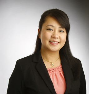 Professional headshot of an Asian woman in black blazer and coral blouse against neutral background.