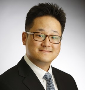 Professional headshot of an Asian man wearing glasses and dark suit against neutral background.